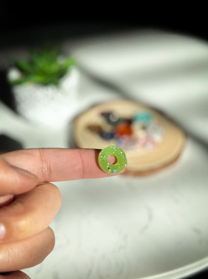 Green donut-shaped earring held between fingers with a blurred background